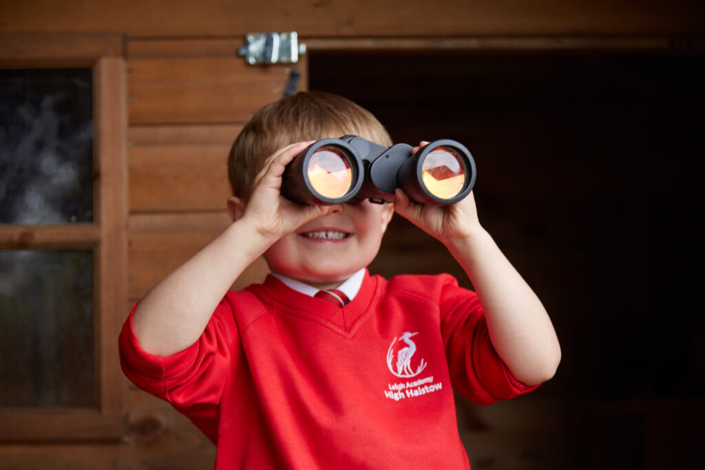A close-up shot of a young child with a wide smile, wearing a dark blue and red school uniform. The child has short, dark hair and is looking directly at the camera. The background is blurred, suggesting an indoor setting with soft, natural light.