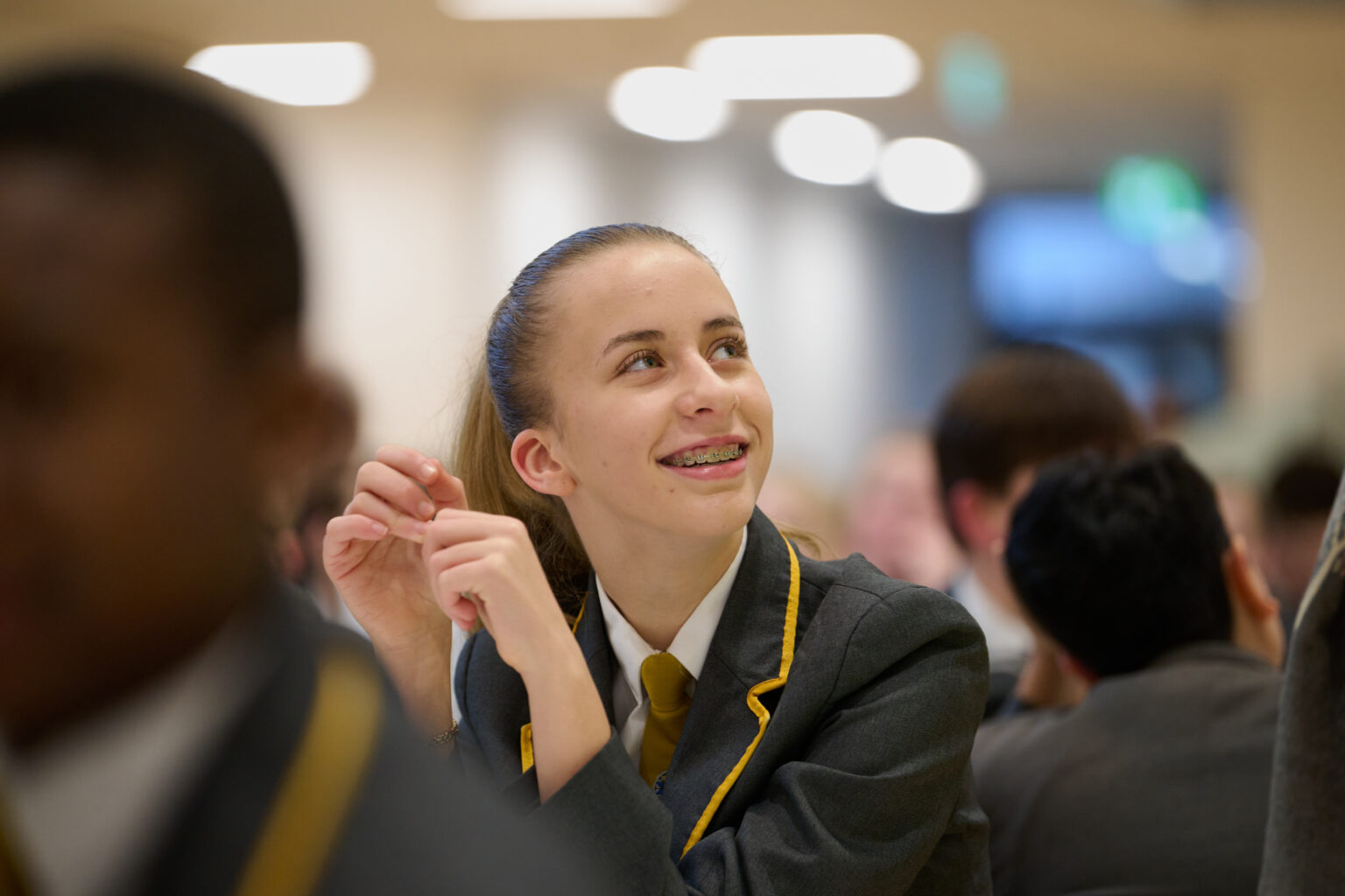 A close-up shot of a smiling student with light brown hair pulled back in a ponytail, wearing a grey school blazer with yellow trim and a yellow tie over a white shirt. The student is looking upwards and to the right, with braces visible on their teeth. The background is blurred, showing other students and warm, circular overhead lights. The focus is on the student's face and upper body.