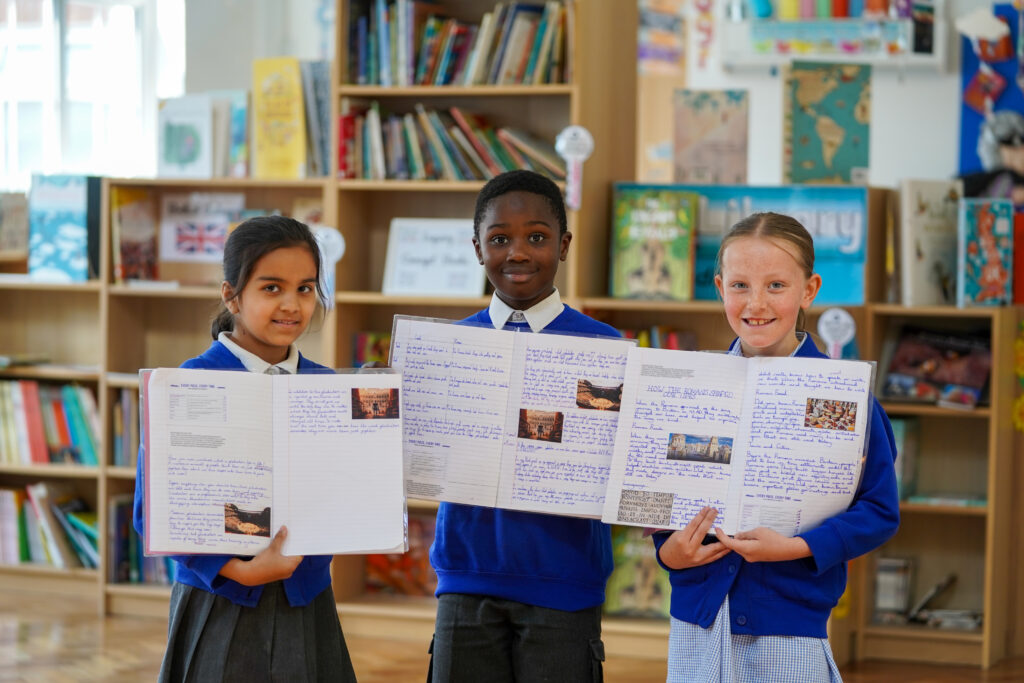 Three children stand in a brightly lit room with bookshelves in the background. On the left, a girl with dark hair and a part in the middle, wearing a white collared shirt and dark grey pleated skirt, holds open a white book with writing and images on it. In the center, a boy with dark skin and short dark hair, wearing a dark blue sweatshirt over a white collared shirt, holds open a larger white book or display board filled with writing and small images. On the right, a girl with light brown hair parted on the side, wearing a dark blue cardigan over a white and grey plaid dress, holds open another white book with writing and images. All three children are looking towards the camera with gentle smiles, holding their books slightly angled for viewing.